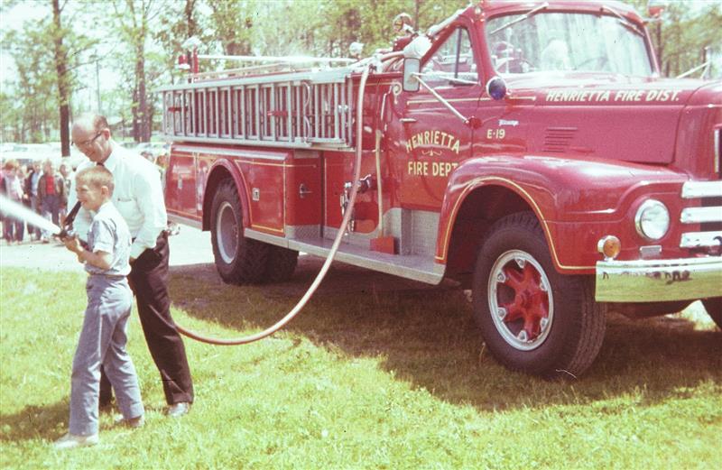 A firefighter and a young student with a fire hose with an o ld fire truck in the background